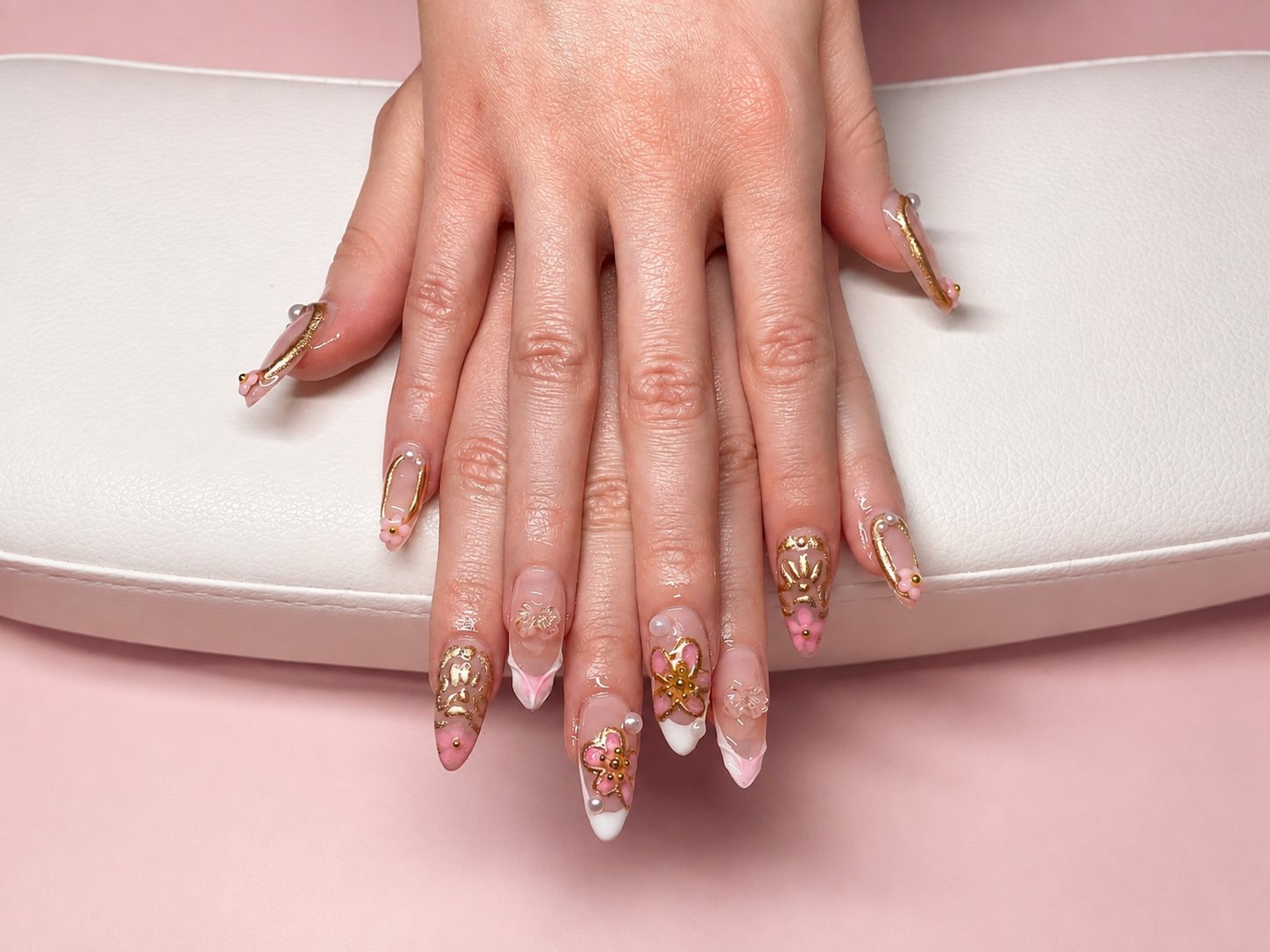 Stiletto GelX set with gold baroque detailing, pink blossoms, and pearls, resting on a white leather cushion